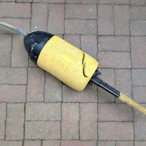 May include: A cylindrical marine buoy featuring a bright yellow central section and black ends. A thick, weathered rope extends from the top, while a black and yellow striped pole protrudes from the bottom. The yellow surface shows signs of wear.