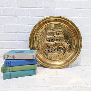 Könnte beinhalten: Eine Vintage-Wanddekoration aus Messing mit einem detaillierten Relief eines Segelschiffs. Die runde Platte hat einen seilartigen Rand. Ein Stapel von vier Büchern mit blauen, grünen und türkisfarbenen Einbänden befindet sich neben der Platte.