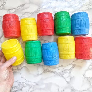 Puede incluir: Un conjunto de diez barriles de juguete de plástico de colores. Los barriles son de varios colores, incluyendo rojo, amarillo, azul y verde. Los barriles están dispuestos en círculo sobre una superficie de mármol blanco.