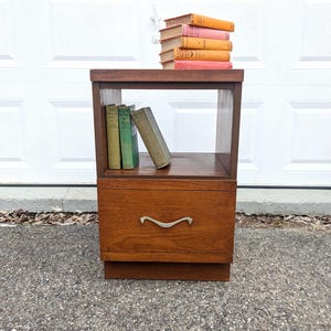 Może przedstawiać: Drewniana szafka nocna z szufladą i otwartą półką. Szafka nocna ma ciepły brązowy kolor i srebrny uchwyt na szufladzie. Na górze ułożono kilka książek, a na półce znajdują się trzy książki.