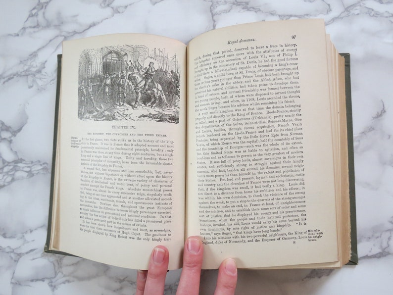 K&ouml;nnte beinhalten: Ein offenes Buch mit einer Schwarzwei&szlig;illustration einer mittelalterlichen Szene. Der Text auf der Seite lautet "Kapitel IV. Das Feudalsystem und der Dritte Stand".