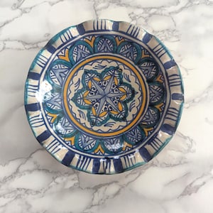 Puede incluir: Un plato decorativo de cerámica con borde ondulado. El plato presenta un diseño central de estrella en azul y amarillo, rodeado de intrincados patrones en colores similares. El borde es azul oscuro con rayas verticales.