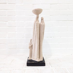 May include: A white ceramic sculpture of three figures, a woman carrying a bowl on her head, a man standing beside her, and a child standing in front of them. The sculpture is on a black base.