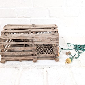 May include: A weathered wooden lobster trap featuring a curved top, slatted sides, and an internal mesh netting compartment. A green nylon rope is attached to the side, ending in a small green and yellow painted wooden buoy float.