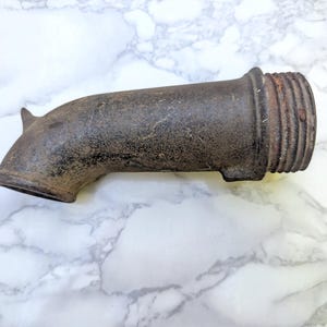 Könnte beinhalten: Ein dunkles, verwittertes Metallrohrstück mit gebogenem Hals und Gewindeende. Die Oberfläche weist Anzeichen von Oxidation und Alter auf, mit einer texturierten, matten Beschaffenheit, sichtbaren Rostflecken und einer kleinen Lasche.