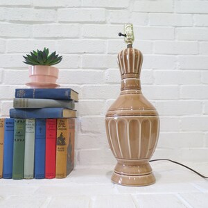 May include: A brown ceramic table lamp with a white base and a white cord. The lamp is on a white brick wall with a stack of books and a small potted plant in front of it.