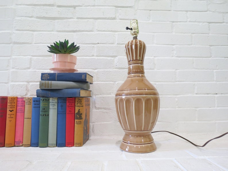 May include: A brown ceramic table lamp with a white base and a white cord. The lamp is on a white brick wall with a stack of books and a small potted plant in front of it.