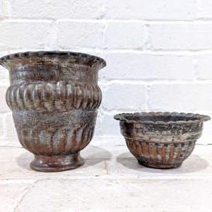 Puede incluir: Dos jardineras antiguas de metal con acabado envejecido. La más grande tiene un cuerpo estriado y un borde decorativo. La más pequeña tiene un borde festoneado. Ambas son de color marrón y plateado.