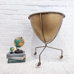 May include: A gold metal planter with a round top and three legs. The planter has a distressed finish and is on wheels. A globe and a stack of books are in the foreground.
