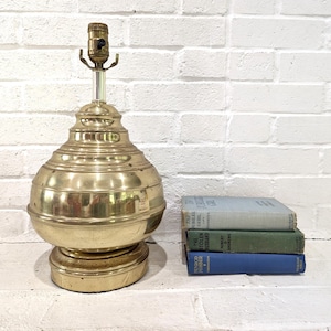 May include: A polished brass table lamp base with a rounded, tiered body and a central metal socket, positioned next to a stack of three hardcover books in light blue, green, and dark blue colors, featuring visible text on the book spines.