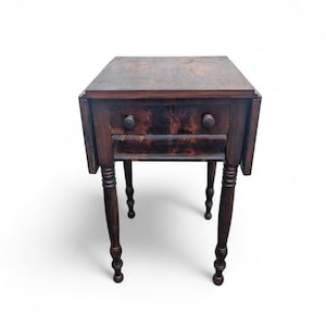 May include: A dark brown antique side table with two drawers and turned legs. The table has a square top with drop leaves and round drawer pulls. The wood grain is visible, giving it a rustic appearance. The table is approximately 61 cm tall.