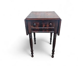 May include: A dark brown antique side table with two drawers and turned legs. The table has a square top with drop leaves and round drawer pulls. The wood grain is visible, giving it a rustic appearance. The table is approximately 24 inches tall.