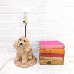 Könnte beinhalten: Eine Keramik-Tischlampe in Form eines goldenen Cocker Spaniels. Die Lampe hat ein braunes Kabel und eine weiße Glühbirne. Die Lampe steht auf einem Holzsockel und befindet sich neben einem Stapel Bücher.