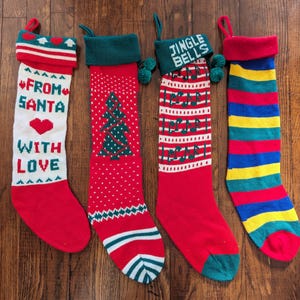 May include: Four Christmas stockings with different designs. One stocking is red and white with the text "From Santa With Love". Another stocking is red and green with a Christmas tree design. A third stocking is red and white with the text "Jingle Bells". The final stocking is red, blue, yellow, and green with stripes.