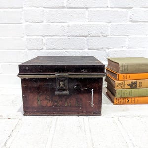 May include: A vintage, dark brown metal trunk with a latch closure, set against a white brick wall. The trunk has a rectangular shape and is slightly distressed. A stack of old books in various colors sits to the right.
