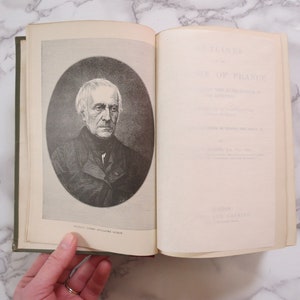 K&ouml;nnte beinhalten: Ein offenes Buch mit einem Schwarzwei&szlig;portr&auml;t eines Mannes auf der linken Seite. Die rechte Seite tr&auml;gt den Titel "Glimpses of France", der in schwarzer Tinte gedruckt ist.