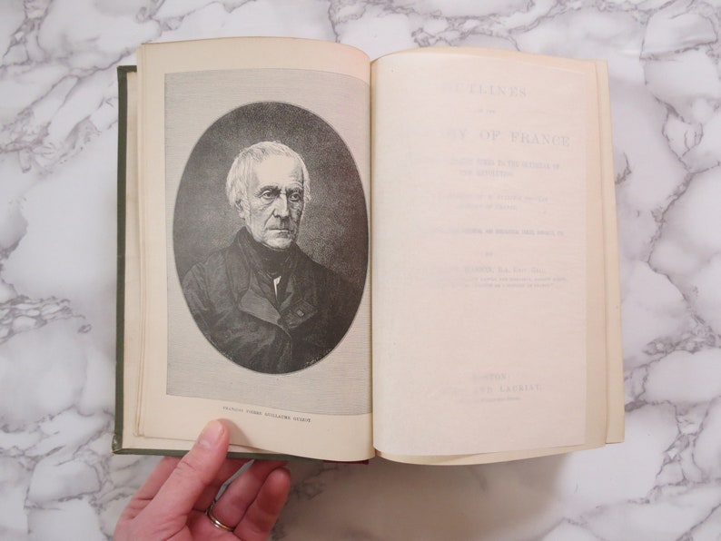 K&ouml;nnte beinhalten: Ein offenes Buch mit einem Schwarzwei&szlig;portr&auml;t eines Mannes auf der linken Seite. Die rechte Seite tr&auml;gt den Titel "Glimpses of France", der in schwarzer Tinte gedruckt ist.
