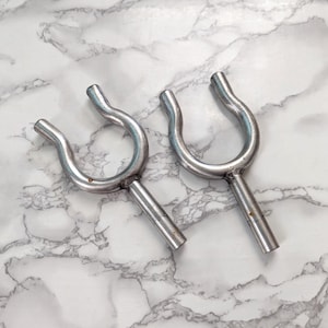 May include: Two silver-colored metal boat cleats, each with a U-shaped top and a cylindrical stem. The cleats are displayed on a white and gray marble-patterned surface. These cleats are designed for securing ropes on boats.