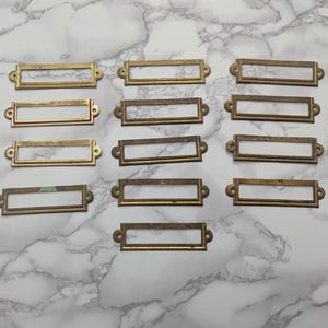 May include: A collection of fourteen rectangular brass label holders arranged in three columns. Each metal frame features a central open slot for inserting paper labels and two small circular holes on the side tabs for mounting with screws or nails.