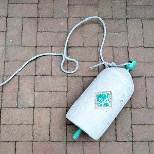 May include: A cylindrical gray marine buoy featuring a central diamond-shaped inset with a weathered green painted surface. The buoy has a green plastic attachment at the top and bottom, with a long, light gray rope tied to the top fixture.