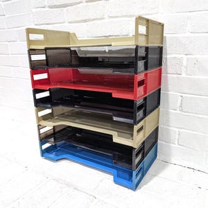 May include: A stack of six modular plastic document trays featuring a vertical design with open sides. The trays are coloured in a repeating sequence of beige, black, red, black, beige, and blue, each with a rectangular cutout handle on the sides.