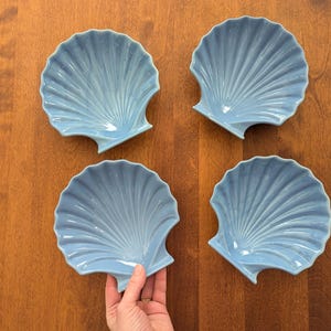 Puede incluir: Cuatro platos en forma de concha de color azul claro sobre una superficie de madera. Los platos tienen una textura acanalada y un acabado brillante. Los platos son de color azul pálido y miden unos 15 cm de diámetro.