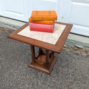 May include: A square wooden side table with a light beige marble tile inlay on the top surface. The table features a central pedestal base with four vertical supports. A stack of four books in orange, yellow, pink, and red sits on the table top.