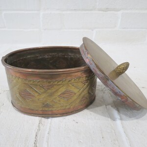 Op de afbeelding: Een ronde, messing en koperen sieradenbox met een decoratief, geweven patroon. De doos heeft een deksel met een kleine messing knop.
