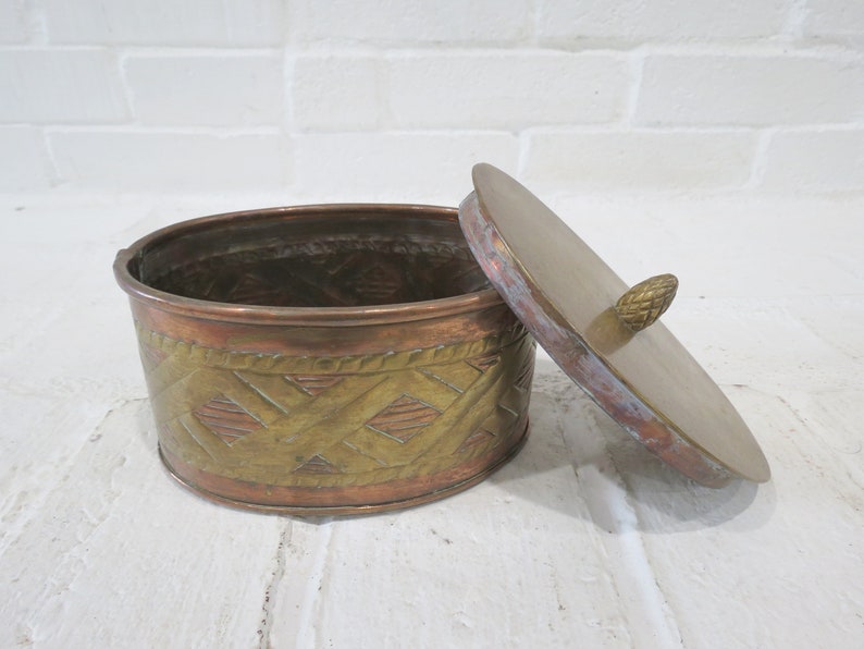 Op de afbeelding: Een ronde, messing en koperen sieradenbox met een decoratief, geweven patroon. De doos heeft een deksel met een kleine messing knop.