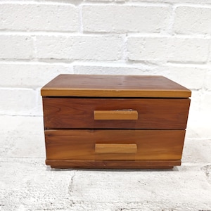 May include: A small wooden chest with two horizontal drawers, each featuring a simple rectangular handle. The unit is constructed from medium-toned wood with a visible grain pattern, showcasing a flat top surface and a base with a slight inset.