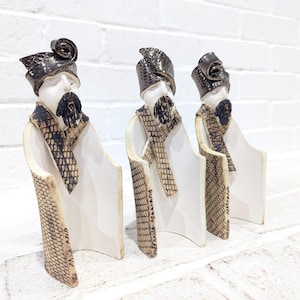 May include: Three white ceramic figurines of people wearing black and brown patterned robes and hats. The figurines are standing in a row on a white surface.