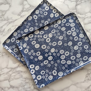 Op de afbeelding: Twee vierkante, marineblauwe serveerbladen met een wit bloemmotief. De bladen zijn versierd met kleine witte bloemen en stippen, waardoor een delicaat, herhalend ontwerp ontstaat. De bladen zijn gemaakt van glanzend materiaal.