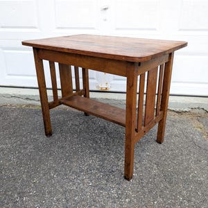 May include: A rectangular wooden table with a medium brown finish, featuring a flat top surface, four straight legs, and a lower shelf positioned between the legs. The side panels of the table are constructed with vertical slats connecting the legs.
