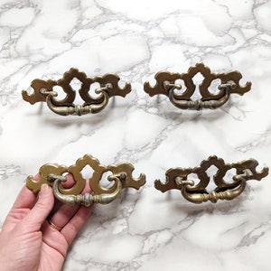 May include: Four antique brass cabinet drawer pulls with ornate designs. Each pull has a curved handle and a decorative backplate.