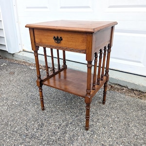 May include: A vintage wooden side table with a drawer and a lower shelf. The table has turned legs and a dark handle on the drawer. The wood has a warm, medium-brown tone. The table is approximately 24 inches tall.