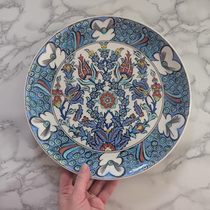 Puede incluir: Un plato de cerámica blanco con un diseño floral azul y rojo. El diseño presenta flores y hojas estilizadas en un estilo turco tradicional.