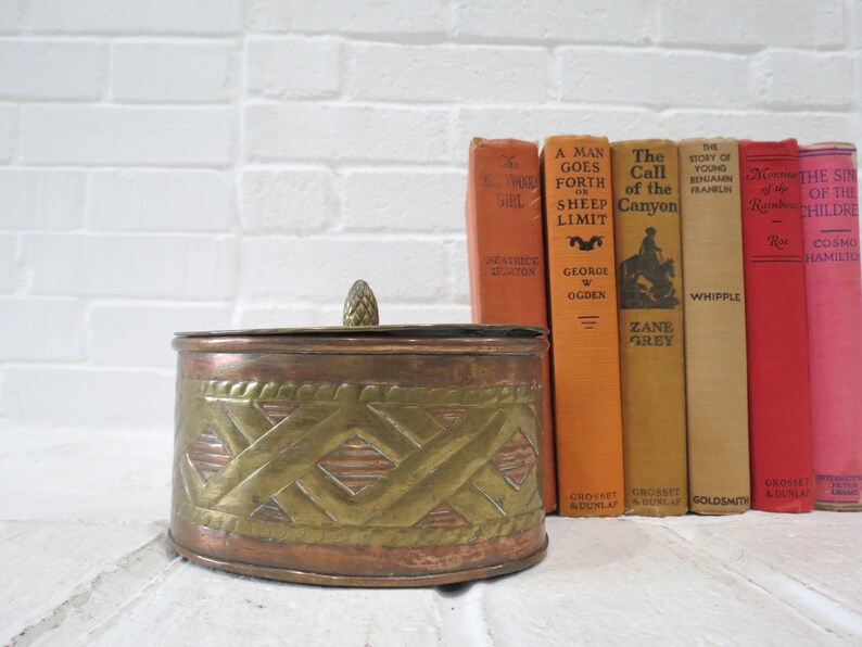 Op de afbeelding: Een ronde koperen en messing sieradenbox met een decoratief geweven ontwerp. De doos heeft een deksel met een klein knopje bovenop. De doos staat voor een stapel vintage boeken.
