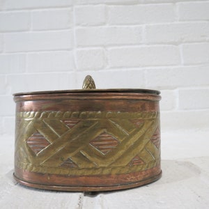 Op de afbeelding: Een ronde koperen en messing sieradenbox met een decoratief geometrisch patroon. De doos heeft een deksel met een kleine knop bovenop.
