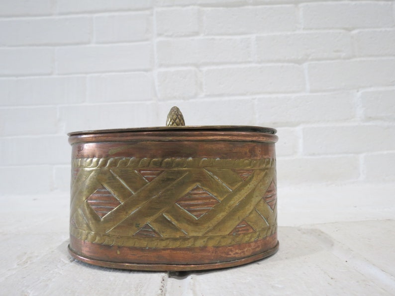 Op de afbeelding: Een ronde koperen en messing sieradenbox met een decoratief geometrisch patroon. De doos heeft een deksel met een kleine knop bovenop.