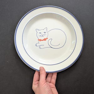 May include: A round ceramic plate with an off-white base and a dark blue rim. The centre of the plate features a simple line drawing of a cat in dark blue ink, accented by a small, bright orange bow tie around the cat's neck.
