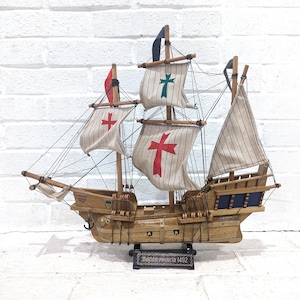 May include: A wooden model of a sailing ship with multiple sails and rigging. The sails are white with red and green crosses. The ship has a brown hull and is labeled "Santa Maria 1492" on a small plaque.