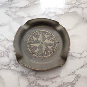 May include: A round, dark gray ceramic tray featuring a central compass rose design with etched cardinal directions N, S, E, and W. The tray has a smooth, matte finish and a slightly raised, scalloped edge with four distinct indentations along the rim.