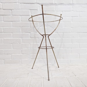 Op de afbeelding: Een metalen plantenstandaard met een ronde bovenkant en drie poten. De standaard is gemaakt van dunne metalen staven en heeft een eenvoudig, modern ontwerp.