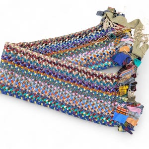 以下が含まれることがあります： 様々な色の布の切れ端で作られた、カラフルな三つ編みのラグ。青、緑、紫、オレンジ色が混ざっています。端には布のタッセルが付いています。このラグはおそらく手作りで、どんな空間にもユニークで質感のある要素を加えています。長さ約120cm。