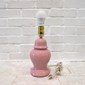 May include: A table lamp with a glossy, light pink ceramic base shaped like a ginger jar. The lamp features a gold-toned metal socket holding a single white LED light bulb. A white electrical cord with a standard two-prong plug extends from the base.
