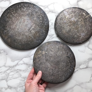 May include: Three antique silver-toned metal plates with ornate floral patterns. The plates are round and vary slightly in size. The plates are arranged on a white and gray marble surface.
