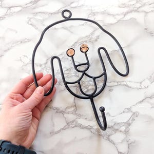 May include: Black metal wall hook in the shape of a dog's face. The hook has a loop at the top for hanging and a single hook at the bottom. The dog's eyes are represented by two small, round, amber-colored beads.