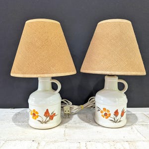 Puede incluir: Un par de lámparas de mesa con bases de cerámica blanca en forma de jarra, con asa y un diseño floral pintado en naranja, amarillo y verde. Cada lámpara tiene una pantalla cónica de tela color canela con textura y cable blanco.