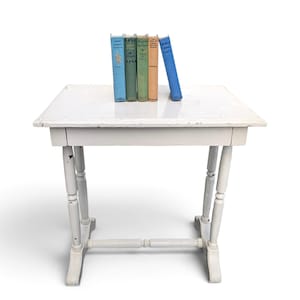 May include: A white painted wooden side table with a rectangular top and four turned legs connected by a horizontal stretcher. Five vintage hardcover books in shades of blue, green, and orange are standing upright on the center of the table surface.
