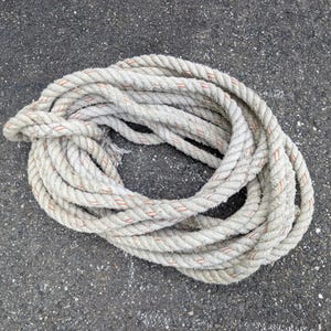 May include: A coiled length of thick, off-white twisted rope featuring thin, intermittent orange tracer threads woven throughout the fibers. The rope is arranged in a circular, overlapping loop pattern, showing a textured, fibrous surface.